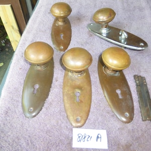 May include: Five antique brass door knobs with keyholes and oval-shaped plates. The knobs are round and have a smooth, polished finish. The plates are slightly curved and have a subtle texture. The knobs are arranged in a row on a light pink surface. The text "8/8/21 A" is written on a white piece of paper below the knobs.