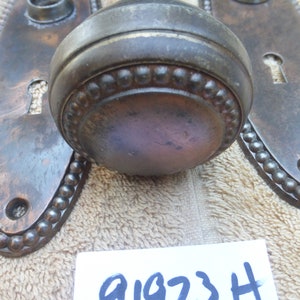 May include: A pair of antique door knobs with oval back plates. The knobs are made of metal and have a beaded edge. The back plates are also metal and have a beaded edge. The knobs are a dark bronze color.