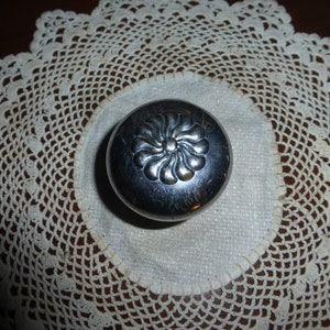 May include: A silver metal trinket box with a floral design on the lid. The box is sitting on a white crocheted doily.