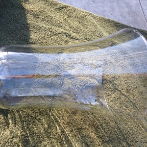 May include: A large, clear glass vase with a rounded bottom and a flared opening. The vase is sitting on a green, textured surface.