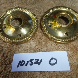 Pode incluir: Dois puxadores de porta dourados com um design de corda ao redor da borda. Os puxadores têm um pequeno furo no centro e três pequenos furos ao redor da borda. Os puxadores estão sobre uma superfície marrom com um pedaço de papel branco com o número 101524 e um círculo nele.