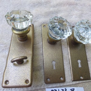 Könnte beinhalten: Drei antike Türklinken mit klaren Glasgriffen und Messingplatten. Die Platten sind goldfarben und haben Schlüssellocher. Die Knöpfe haben alle unterschiedliche Formen.