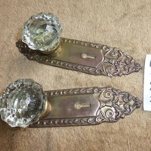 May include: Two door knobs with clear, faceted glass handles attached to rectangular brass backplates. Each backplate features an ornate, embossed scrollwork pattern and a central keyhole. A white paper tag with the handwritten text 41024 I is present.
