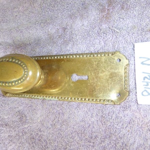 May include: Brass door knob and backplate with a decorative beaded border. The backplate has a keyhole and two small holes. The knob is oval shaped with a beaded border.