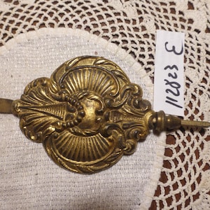 May include: Ornate, antique brass clock pendulum bob with a detailed, symmetrical design. The decorative piece features a central face-like motif, radiating lines, and scrollwork. A small tag with the number "112823 E" is visible.