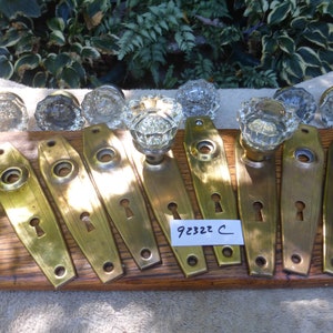 May include: Set of eight antique brass door knobs and backplates with clear glass knobs. The backplates are oval shaped with a keyhole. The knobs have a faceted design. The set is labeled "92322 C".