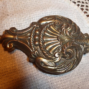 May include: Ornate, antique brass-colored decorative object with intricate floral and scrollwork details. The object has a central, fan-like design with a rope-like border and a rod extending from each end. The piece is likely a decorative element.