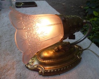 May include: An antique table lamp with a frosted pink glass shade featuring an etched floral design. The lamp has a brass-colored base and a curved arm. The lamp is lit, emitting a warm glow.