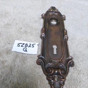 Puede incluir: Placa de puerta de bronce ornamentada con un ojo de cerradura y adornos de volutas. La placa tiene forma rectangular con un agujero circular en la parte superior y el texto "52825 G" en una etiqueta blanca.