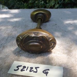 May include: Antique brass door knob with an oval, ornate design. The knob has a long, slender stem connecting two rounded ends. The image includes a label with the numbers "52125 G".