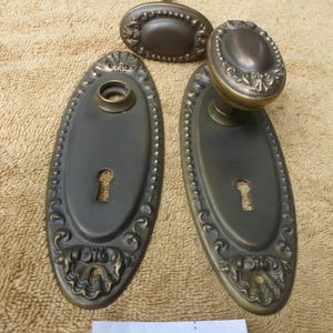 May include: Antique bronze door hardware set. The set includes two oval backplates with keyholes, a doorknob, and a small oval piece. The hardware has ornate detailing and a vintage aesthetic. The number "82825 E" is printed on a white label.