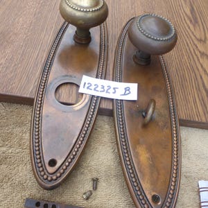 Könnte beinhalten: Zwei antike Türknöpfe aus Bronze mit ovalen Rückplatten. Die Rückplatten haben eine perlenbesetzte Umrandung und einen runden Knauf. Das Bild enthält eine kleine Metallstange, Schrauben und ein Etikett mit dem Text "122325 B".