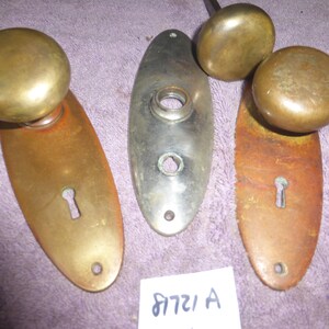 May include: Three antique brass door knobs with oval escutcheons. The escutcheons have keyholes and are in various states of wear. The knobs are round and have a smooth finish.