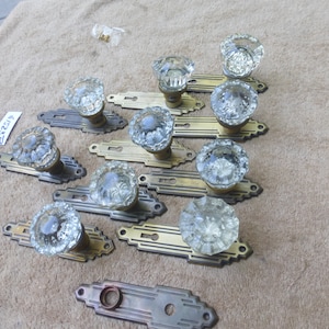 Puede incluir: Conjunto de diez pomos de puerta antiguos con vidrio transparente y placas de latón. Los pomos tienen un diseño facetado y las placas tienen una cerradura y detalles decorativos. Las placas están etiquetadas con el número 23219.