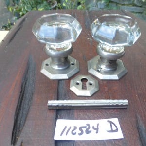 May include: Two antique glass door knobs with octagonal bases and a silver finish. The knobs are on a wooden surface with a metal screw and a keyhole plate.