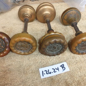 May include: Four antique brass door knobs with ornate floral designs. The knobs are in various stages of wear and patina. The knobs are arranged in a row on a brown surface. The text "1-26.24 B" is written on a white label.