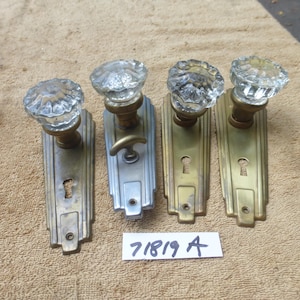 Könnte beinhalten: Vier antike Türknöpfe mit klaren, facettierten Glasgriffen und messingfarbenen Rückplatten. Ein Knopf hat eine silberfarbene Rückplatte und einen Griff. Die Rückplatten haben Schlüssellöcher und ein dekoratives Design. Ein Etikett lautet "71819 A".