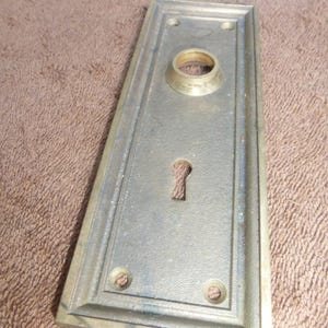 Może przedstawiać: Prostokątna mosiężna płyta drzwiowa z otworem na klucz i okrągłym otworem na klamkę. Płyta ma podniesioną krawędź i trzy otwory na śruby. Tekst "92823 V" znajduje się na białej etykiecie w tle.