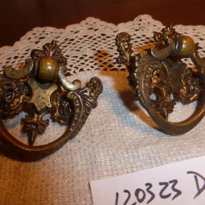 May include: Two ornate brass drawer pulls with a decorative floral design. The pulls have a ring shape and a round knob in the center.