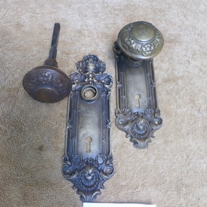 May include: Two antique brass door knobs with matching ornate rectangular backplates featuring intricate scrollwork and keyholes. One knob is detached with a square spindle, while the other is attached to its plate. A white tag displays the text 72224C.