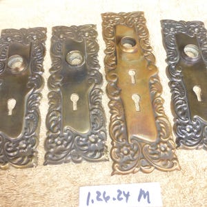 May include: Four antique brass door plates with ornate floral scrollwork. Each plate features a keyhole and a circular opening for a doorknob. The plates vary in color from dark bronze to a lighter brass.