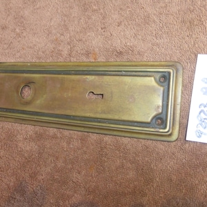 May include: A rectangular brass door plate with a keyhole and a circular hole. The plate has a decorative border and is slightly tarnished. A small white label with the numbers "92823 BA" is next to the plate.