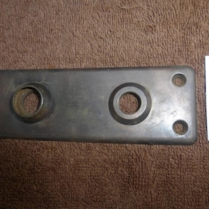 Puede incluir: Una placa metálica rectangular con dos aberturas circulares y cuatro orificios para tornillos. La placa tiene un acabado plateado con algo de desgaste. Una etiqueta blanca con el texto "92883 R" está en la imagen.