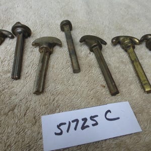 Puede incluir: Un conjunto de herrajes metálicos antiguos, posiblemente tiradores o pomos, con varios diseños de cabeza y vástagos roscados. Las piezas están envejecidas con un acabado bronceado. Una etiqueta blanca con el número "51725 C" es visible.