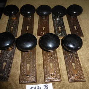 Puede incluir: Una colección de pomos de puerta antiguos con manijas redondas negras y placas traseras ornamentadas de color bronce. Las placas traseras presentan un ojo de cerradura y patrones decorativos. Los pomos están dispuestos sobre una superficie marrón.