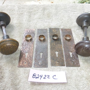 Puede incluir: Cuatro placas de puerta de latón antiguo con diseños ornamentados y agujeros para llaves, y dos pomos de puerta de latón antiguo a juego con acentos negros. Las placas y los pomos están dispuestos sobre una superficie peluda de color marrón claro. El texto "82922 C" está escrito en un trozo de papel blanco.