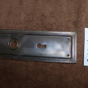 Könnte beinhalten: Rechteckige antike Türplatte mit Schlüsselloch und kreisförmiger Öffnung. Die Platte besteht aus dunklem Metall mit dekorativem Rand. Zwei kleine Löcher sind in der Nähe der Ecken sichtbar. Ein weißes Etikett mit der Nummer "92803 U" befindet sich rechts.