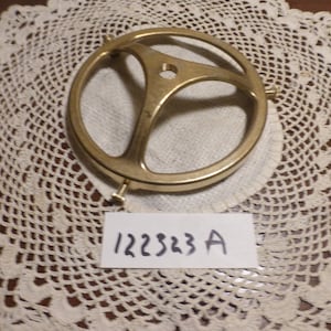 Può includere: Un portalampada in metallo dorato con tre punte. Il portalampada è rotondo e ha una superficie liscia. Il testo "122383 A" è scritto su un pezzo di carta bianco.