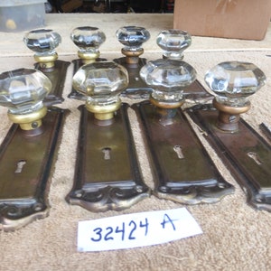 May include: Set of eight antique door knobs with clear glass knobs and brass plates. The plates have a keyhole and decorative detailing. The knobs are arranged on a brown surface with a white tag that reads "32424 A".