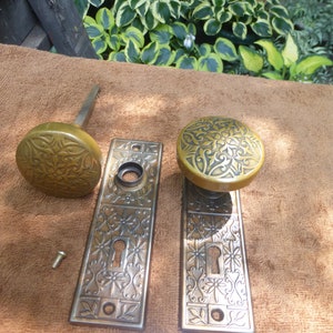 May include: Two antique brass door knobs with ornate floral designs and keyholes. The knobs are attached to rectangular plates with matching designs. The plates are on a brown surface with the number 52423D written on a white piece of paper.