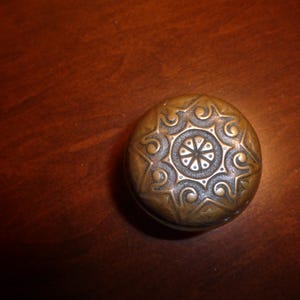 May include: A round, antique bronze cabinet knob with an ornate, symmetrical design. The knob features a central circular pattern surrounded by stylized floral or star-like elements. The knob is set against a dark wood surface.