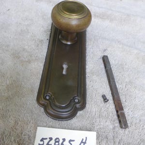May include: Antique brass door knob set with a keyhole and a decorative backplate. Includes a separate metal piece and a screw. The set is aged with a vintage look. A tag with the text "52825 H" is also present.