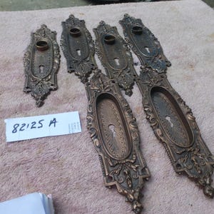 Może przedstawiać: Kolekcja antycznych okuć do drzwi z brązu, w tym ozdobne rozety i płyty uchwytów. Elementy charakteryzują się szczegółowymi wzorami kwiatowymi i przewijanymi, z otworami na klucze i okrągłymi mocowaniami uchwytów. Tekst "82125 A" jest widoczny.