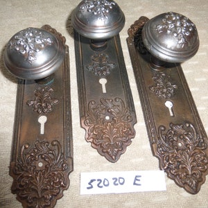 以下が含まれることがあります： 装飾的なデザインと銀色の塗装仕上げが施されたアンティークなドアノブが3つ。ノブは、複雑な花柄が施された長方形のプレートに取り付けられています。プレートは、ダークブラウンの色で、使い古した仕上げになっています。ノブの下の白いタグには、「520 20 E」という文字が書かれています。