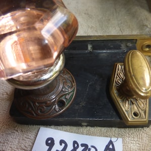 May include: Antique door hardware set featuring a faceted, rose-colored glass doorknob, a decorative bronze knob, and a black metal plate. The set includes a bronze strike plate and a decorative bronze backplate.