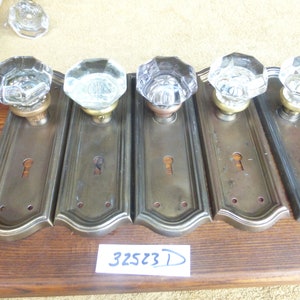 Könnte beinhalten: Fünf antike Messing-Türknäufe mit klaren, achteckigen Glasknöpfen. Die Knäufe sind auf rechteckigen Platten mit einem Schlüsselloch und dekorativen Kanten montiert. Die Platten sind in einer Reihe auf einer Holzoberfläche angeordnet. Die Nummer 325231 ist auf die Oberfläche geschrieben.