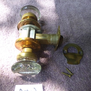 May include: A brass door knob set with clear glass knobs. The set includes a door knob, a strike plate, and two screws.