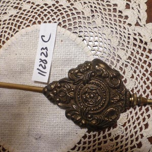 May include: An antique brass clock pendulum with an ornate, floral design. The pendulum has a long, slender rod and a decorative, detailed face. A white tag with the numbers "112823 C" is attached.