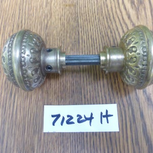 May include: A pair of round brass door knobs with an embossed decorative pattern connected by a central metal spindle. A white rectangular paper tag is positioned below the knobs with the handwritten text 71224 H written in blue ink.
