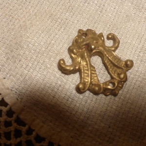 May include: A gold-colored, ornate metal escutcheon with a keyhole shape. The decorative piece features swirling, curved designs and is set against a textured, off-white fabric.