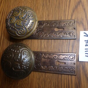 May include: Two matching brass door knobs with circular plates featuring intricate embossed scrollwork and leaf patterns. Each knob is attached to a rectangular backplate with a keyhole cutout. A small white paper tag displays the text 41124 K.