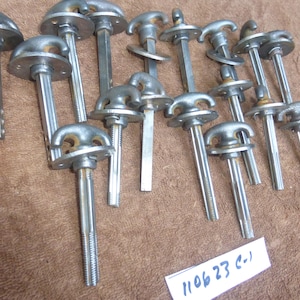 May include: A collection of 14 silver metal door handles with a unique design. Each handle features a round base with a threaded shaft and a decorative top with two loops. The handles are arranged in a random pattern on a brown surface. The text "11 06 23 C-1" is visible in the lower right corner of the image.