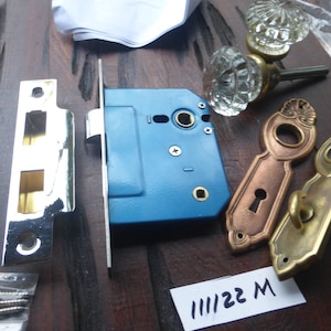 May include: A set of antique door hardware in brass and chrome. The set includes a door knob, a door plate, a door latch, and screws. The door knob is clear glass with a faceted design. The door plate is brass with a keyhole and a decorative design. The door latch is chrome with a blue finish. The screws are silver.