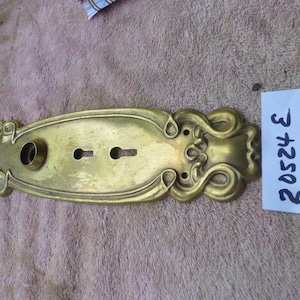 May include: A vintage brass door plate with ornate scrollwork and keyholes. The plate has a tarnished finish, showing signs of age. A white label with the number "20524 E" is attached.