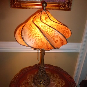 May include: An ornate table lamp with a unique, ruffled lampshade. The shade is a warm orange color with brown decorative patterns. The lamp has a detailed, bronze-colored base and a pull chain. The lamp is sitting on a decorative, round table.