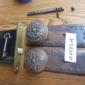 Könnte beinhalten: Antikes Türbeschlagsset, bestehend aus einem schwarzen Schlosskasten mit Schlüssel, einem Messing-Schließblech und zwei verzierten Türgriffplatten aus Bronze. Enthält eine kleine Metallstange und Schrauben. Die Platten haben ein florales Design und die Nummer "120923 A" auf einem Aufkleber.